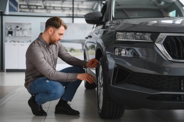 Adam araba galerisindeki yeni bir aracın lastiklerini kontrol ediyor, satın almadan önce karar veriyor.