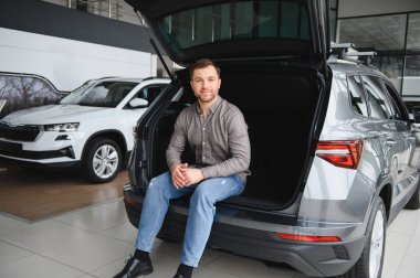 Bir otomobil galerisindeki deneyimin tadını çıkarırken bir arabanın bagajında oturan mutlu bir adam.