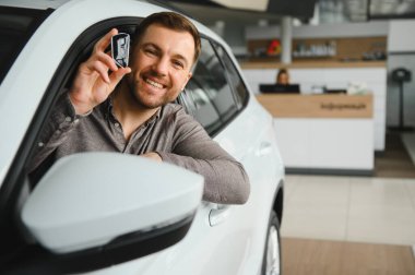 Hevesli bir adam araba anahtarı tutuyor, modern bir araba galerisinde yeni bir aracın yanında duruyor, başarılı bir satın alma olduğunu gösteriyor.