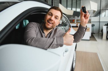 Araba anahtarlarını ve baş parmaklarını gösteren mutlu bir adam, bir galeride araba camından sarkıyor, yeni alımını kutluyor.