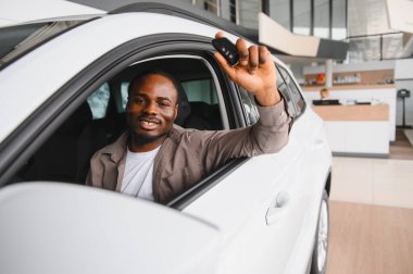 Gülümseyen adam galeride yeni bir arabada oturuyor, gururla araba anahtarlarını gösteriyor ve satın almanın heyecanıyla eğleniyor.