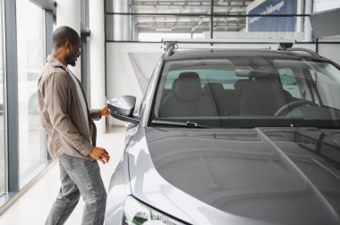 Parlak bir otomobil galerisinin içinde heyecanlı ve tatmin olmuş bir şekilde dikiz aynasını kontrol eden mutlu bir müşteri.