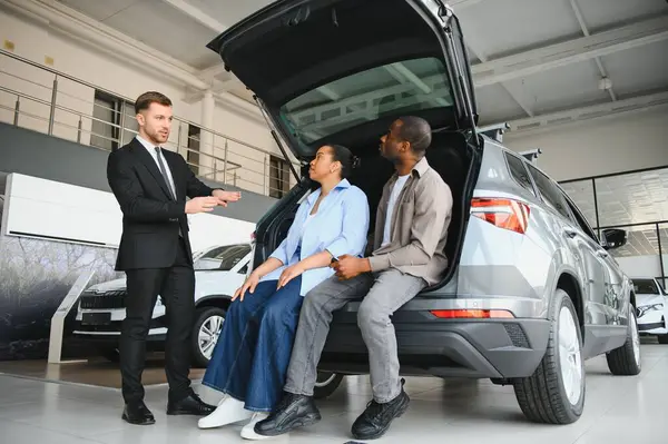 Araba satıcısı, bir galeride açık bagajda oturan bir çifte yeni arabanın özelliklerini sunuyor.