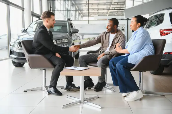 Satıcı, Afro-Amerikan çiftiyle el sıkışıyor. Modern bir bayiliğin içinde yeni bir araba için anlaşma yapıyor.