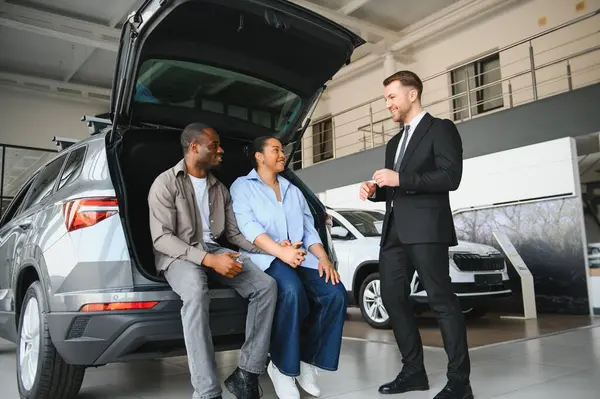 Satış elemanı modern bayilikte özelliklerini anlatırken mutlu çift yeni arabanın bagajında oturuyor.