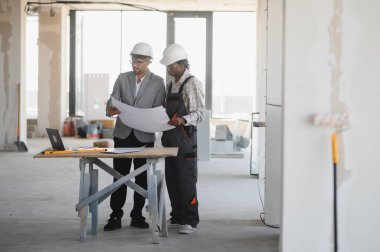 Mimar ve müteahhit şapka takıyor ve inşaat alanının planlarını inceliyor, bina planlarını tartışıyor.