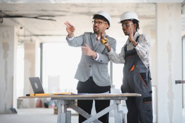 Arap mimar ve Afrikalı Amerikalı müteahhit inşaat sahasında planlarını tartışıyor, inşaat sektöründe takım çalışması ve çeşitliliği sergiliyor