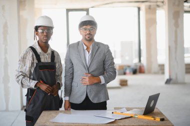 Güvenlik kaskı takan mimar ve müteahhitler, hareketli bir inşaat sahasının ayrıntılı planlarını gözden geçirip proje üzerinde işbirliği yapıyorlar.