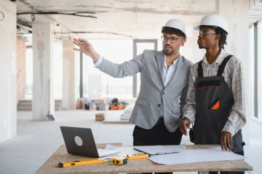 İnşaat planları üzerinde işbirliği yapan mimar ve inşaatçı, inşaat alanındaki planları incelerken proje ayrıntılarını tartışıyor