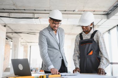 Mimar ve müteahhit başlık takıyor, hareketli bir inşaat alanının planlarını dikkatlice gözden geçiriyor, proje planları üzerinde işbirliği yapıyor.