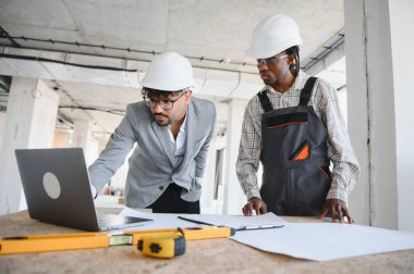 Şapkalı mimar ve müteahhit planları inceliyor ve inşaat alanında dizüstü bilgisayar kullanıyor.