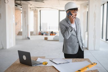 Arap mimar takım elbise giyip, akıllı telefondan konuşurken inşaat sahasında elinde planlar ve dizüstü bilgisayarla masada dikiliyor.