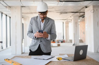 İnşaat alanında takım elbise ve kask giyen Arap mimar akıllı telefon kullanıyor, projeyi yönetiyor ve e-postaları kontrol ediyor