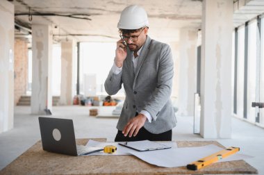 Arap mimar takım elbise giyiyor ve inşaat alanını yönetiyor, telefonla konuşuyor ve planları kontrol ediyor.