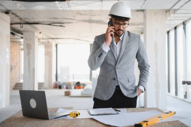 İnşaat alanında akıllı telefon kullanan takım elbiseli ve kasklı bir Arap mimar, masanın üzerinde planlar ve dizüstü bilgisayar var.