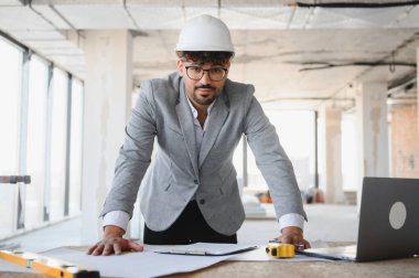 Takım elbiseli ve şapkalı Arap mimar hareketli bir inşaat sahasının detaylı bina planlarını inceliyor ve projenin hassasiyetini sağlıyordu.