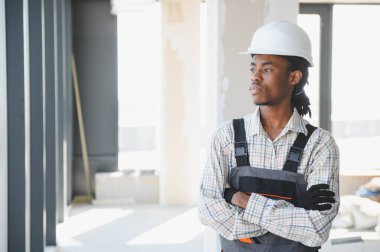 Güvenilir Afro-Amerikan inşaat işçisi güvenlik kaskı takıyor ve kolları bağlı inşaat alanını inceliyor.