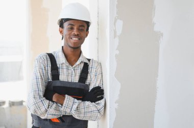 Genç Afro-Amerikan inşaat işçisi kollarını kavuşturup gülümsüyor, iş tulumu ve şapka giyiyor, kısmen sıvanmış bir duvarın yanında duruyor.
