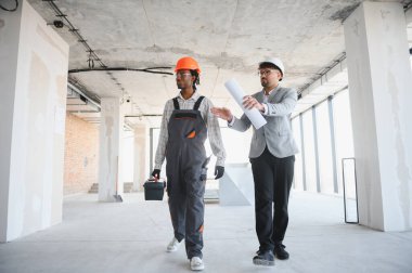 Mimar ve inşaatçı kısmen inşa edilmiş bir binanın planlarını tartışıyor, inşaat sektöründe takım çalışması ve çeşitliliği sergiliyor