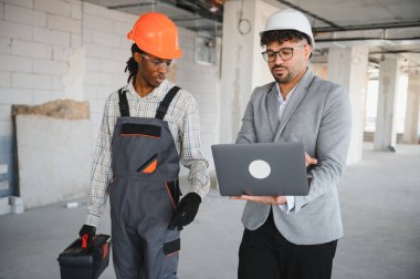 İnşaat alanı ziyareti sırasında inşaat planlarını gösteren, tulum giyen ve alet çantası taşıyan bir mimar