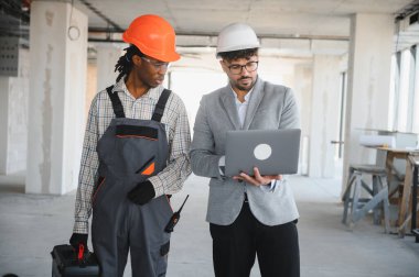 Mimarın elinde dizüstü bilgisayar var. İnşaat aşamasında olan bina planlarını gösteriyor.