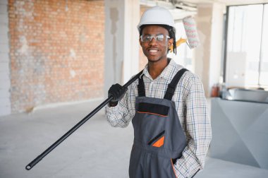 Afrikalı Amerikalı inşaat işçisi gülümsüyor. Omzunda teleskopik bir boya silindiri taşıyor. Güvenlik gözlüğü ve miğfer takıyor.