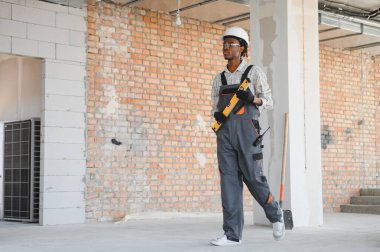 Genç Afro-Amerikan inşaat işçisi inşaat sahasında yürürken ruh seviyesinde üniforma ve şapka giyiyor.