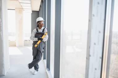 Genç Afro-Amerikan inşaat işçisi elinde bir aletle inşaat halindeki bir binanın penceresinden dışarı bakıyor.