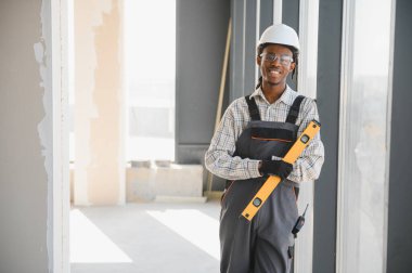 Gülümseyen Afro-Amerikan inşaat işçisi. Güvenlik kaskı takıyor. İnşaat halindeki bir binanın içinde ruh seviyesini tutuyor.