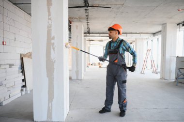 İnşaat işçisi uzun saplı bir boya rulosu kullanıyor. İnşaat halindeki bir binanın duvarlarına astar kullanıyor.