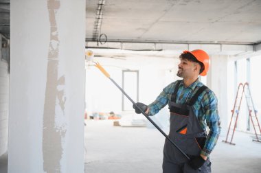 Arap müteahhit tulum giyiyor ve inşaat alanında uzun bir fırça kullanarak duvar boyuyor.