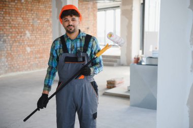 Gri üniformalı ve şapkalı Arap bir ressam, elinde boya silindiri tutarken bir inşaat alanında çalışıyor.