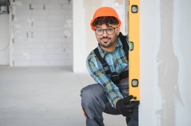 İnşaat aşamasındaki yeni bir binanın ruh seviyesini kullanarak duvarın dikeyliğini kontrol eden genç bir inşaat işçisi.