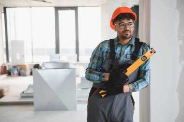 Arap mühendis koruyucu iş giysisi giyiyor ve inşaat halindeki bir binanın içinde bir araç tutuyor, başka tarafa bakıyor.