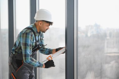 Arap inşaat işçisi, modern yüksek katlı bir binanın pencere çerçevesine dolgu macunu sürüyor, hava geçirmez ve hava geçirmez kurulumunu sağlıyor.