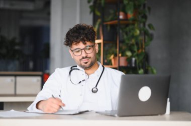 Laboratuvar önlüğü ve steteskop giyen odaklanmış erkek pratisyen modern bir ofiste dizüstü bilgisayarı üzerinde çalışırken not alıyor.