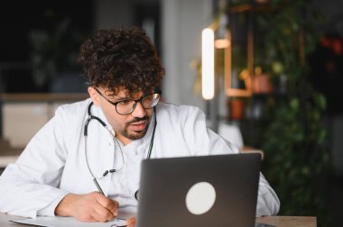 Laboratuvar önlüğü ve steteskop giyen odaklanmış erkek doktor, laptop kullanıyor ve çağdaş ofiste online danışmanlık veya tıbbi araştırma sırasında not alıyor