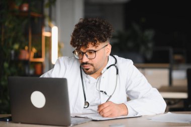 Laboratuvar önlüğü ve steteskop giyen odaklanmış erkek doktor dizüstü bilgisayar kullanıyor ve not alıyor, bitkilerle modern ofiste çalışıyor.