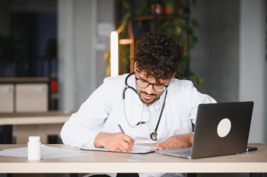 Laboratuvar önlüğü ve steteskop giyen odaklanmış erkek doktor, modern tıp bürosunda dizüstü bilgisayar kullanırken panoya notlar yazıyor.
