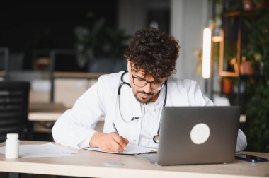 Odaklanmış erkek doktor not alıyor ve modern bir tıp ofisinde dizüstü bilgisayar kullanıyor, profesyonelliği ve hasta bakımına olan bağlılığını gösteriyor