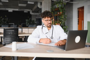 Laboratuvar önlüğü ve steteskop giyen erkek doktor laptopunu kullanıyor ve revirde çalışırken not alıyor.