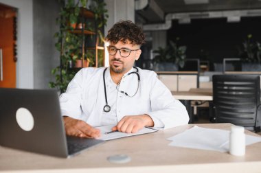 Laboratuvar önlüğü ve steteskop giyen odaklanmış erkek doktor dizüstü bilgisayar kullanıyor, çağdaş klinikte tıbbi kayıtlarla çalışıyor.