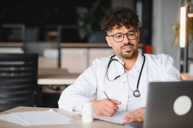 Laboratuvar önlüğü ve steteskop giyen odaklanmış erkek doktor, tıp bürosunda masada oturuyor, dizüstü bilgisayar kullanıyor ve not alıyor, online danışmanlık yapıyor veya hasta bilgilerini araştırıyor.