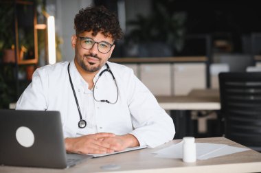 Kendine güvenen Arap erkek doktor modern klinikte dizüstü bilgisayarla çalışıyor, online danışmanlık yapıyor ya da tıbbi bilgi araştırıyor.