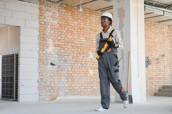 Genç Afro-Amerikan inşaat işçisi inşaat sahasında yürürken ruh seviyesinde üniforma ve şapka giyiyor.