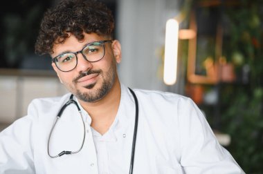 Gözlüklü, beyaz önlüklü, steteskoplu, gülümseyen ve kameraya bakan genç bir Arap erkek doktorun portresi.