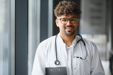 Kendine güvenen bir Arap erkek doktorun portresi gülümseyen ve elinde bir pano tutan, bir hastane ortamında uzmanlık ve profesyonellik gösteren