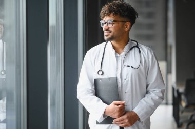 Beyaz önlüklü ve steteskoplu güvenilir Arap erkek doktor modern hastanenin penceresinden dışarı bakıyor.