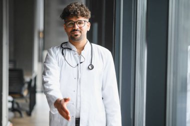Dost canlısı Arap doktor el sıkışmak için elini uzatıyor, modern hastane koridorunda hastayı karşılıyor.