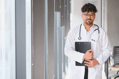 Beyaz önlüklü ve steteskoplu güvenilir Arap erkek doktor modern hastane koridorunda dururken elinde pano tutuyor.